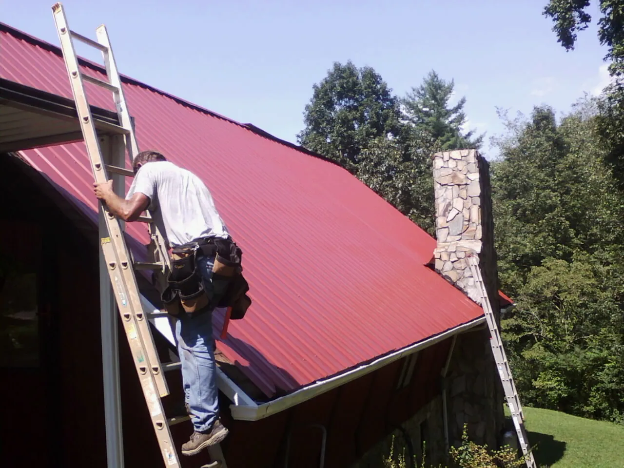 Roofer on a ladder beside a red metal roof and stone chimney — Soffit & Fascia Repair by Roofing Brothers in Mundelein